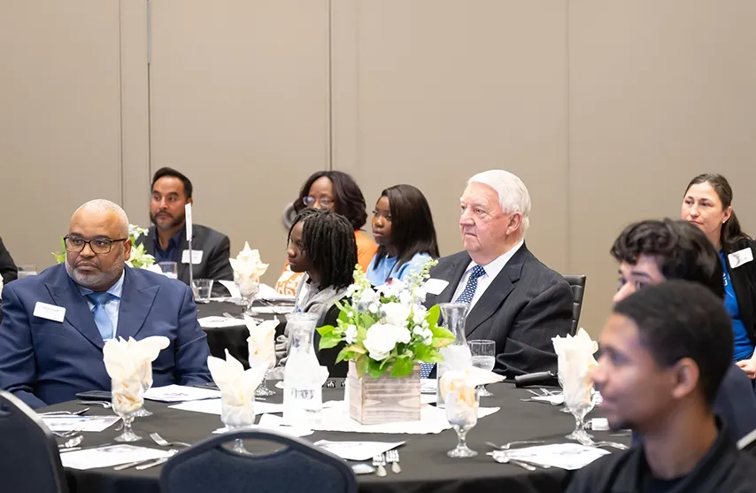 A group of formally dressed people sit at a table, listening to a speech.