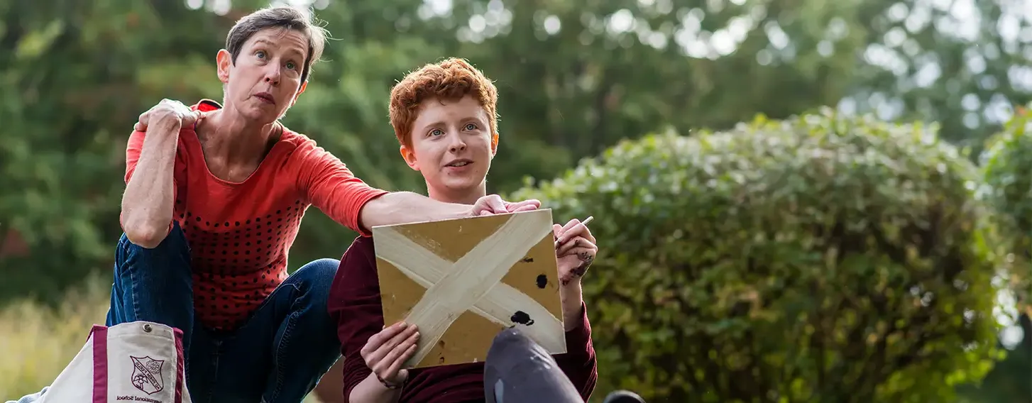 A woman sitting on the grass with a student who is holding a painting.